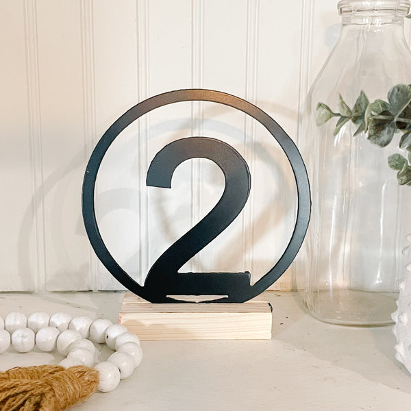 Decorative number '2' on a wooden stand with a white bead necklace and glass jar in the background.