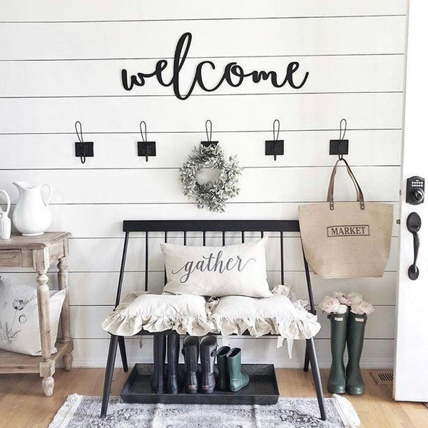 Welcome sign above a bench with decorative pillows and boots in a home entryway.