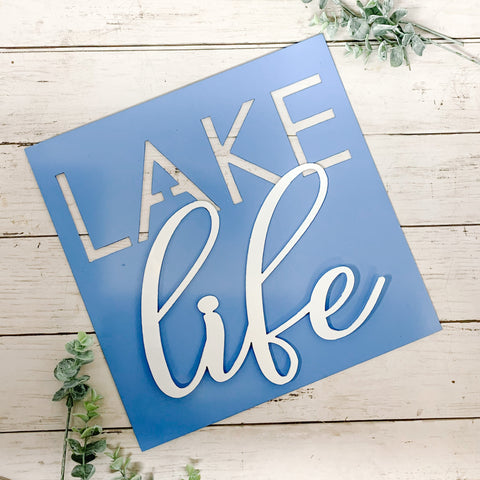 A blue metal sign with the words 'LAKE life' in white script, placed on a wooden surface with greenery to the side.